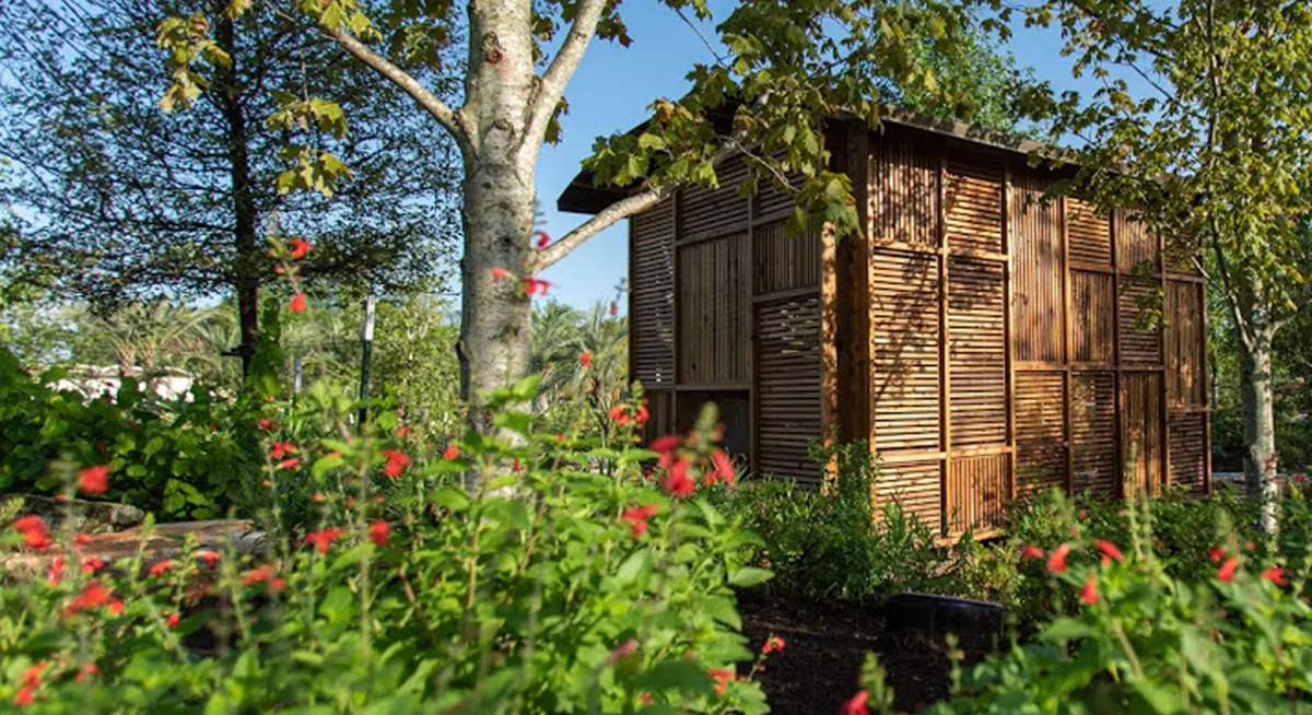 Wooden structure nestled among lush greenery and red flowering plants at the Houston Botanic Garden, crafted with intricate wood paneling by Renfrow+Co Wooden structure nestled among lush greenery and red flowering plants at the Houston Botanic Garden, crafted with intricate wood paneling by Renfrow+Co