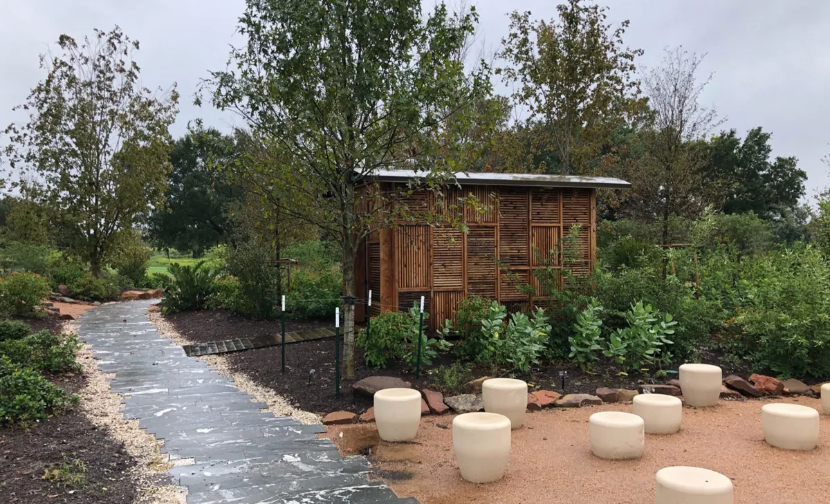 Wooden slat structure in a natural setting at Houston Botanic Garden, featuring white stone stools on a path and greenery in the background Wooden slat structure in a natural setting at Houston Botanic Garden, featuring white stone stools on a path and greenery in the background