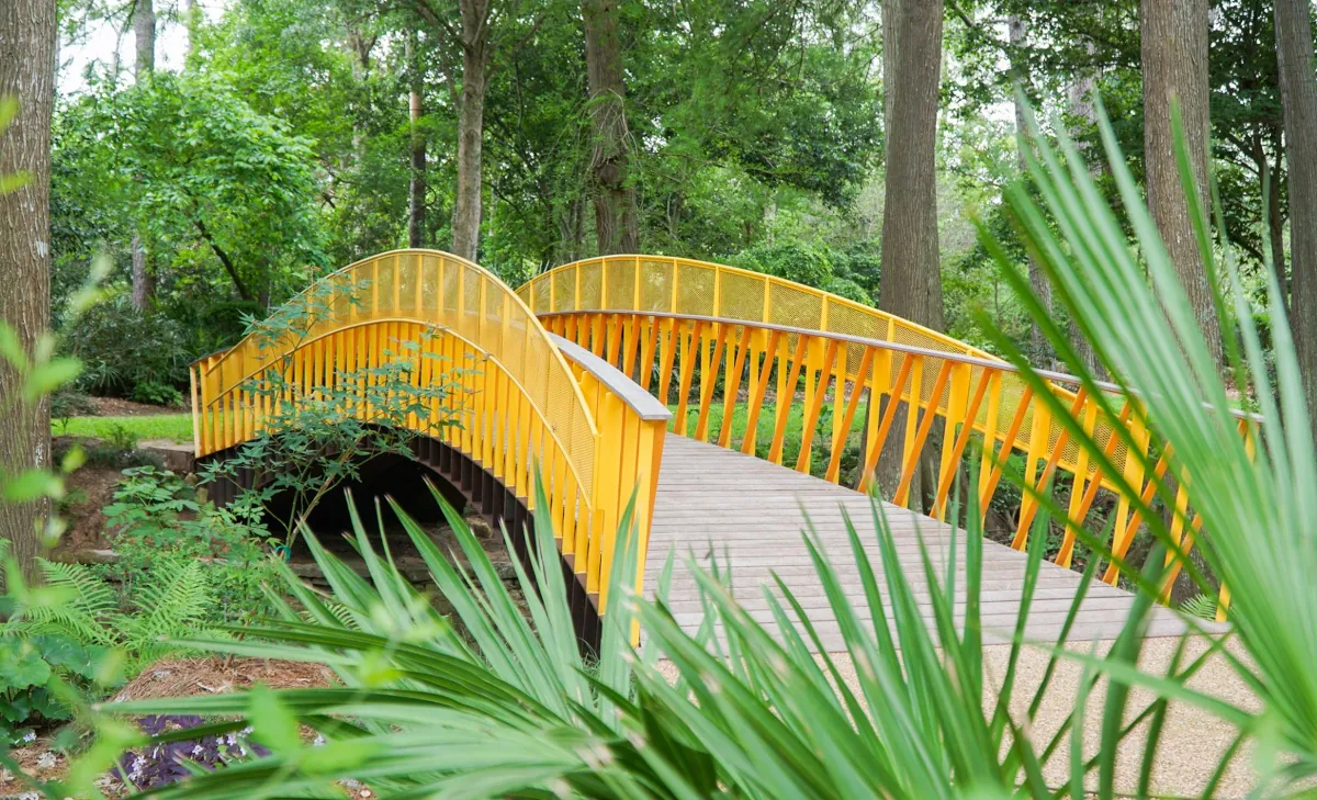 A vibrant yellow bridge, designed by Renfrow+Co, with a curved path, metal mesh panels, and slanted beams, creating a striking contrast in the landscape of John Fairey Garden